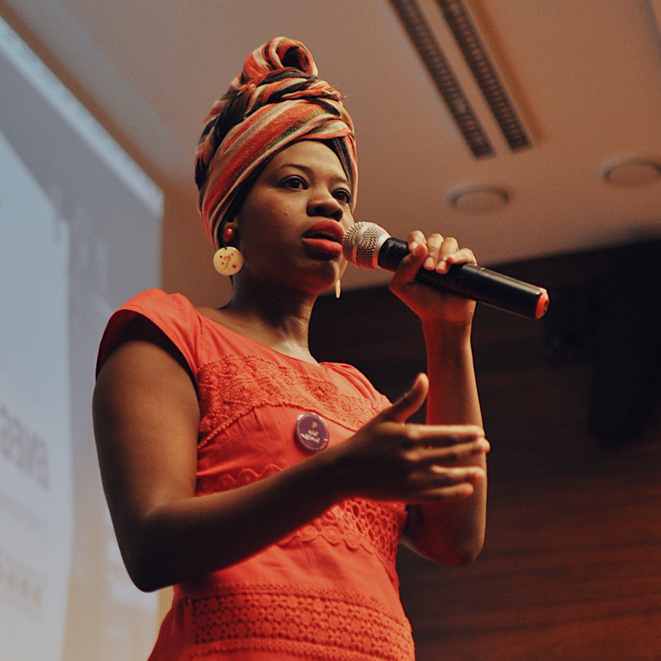 Mulher negra vestindo turbante e vestido laranja, na mão esquerda segura um microfone
