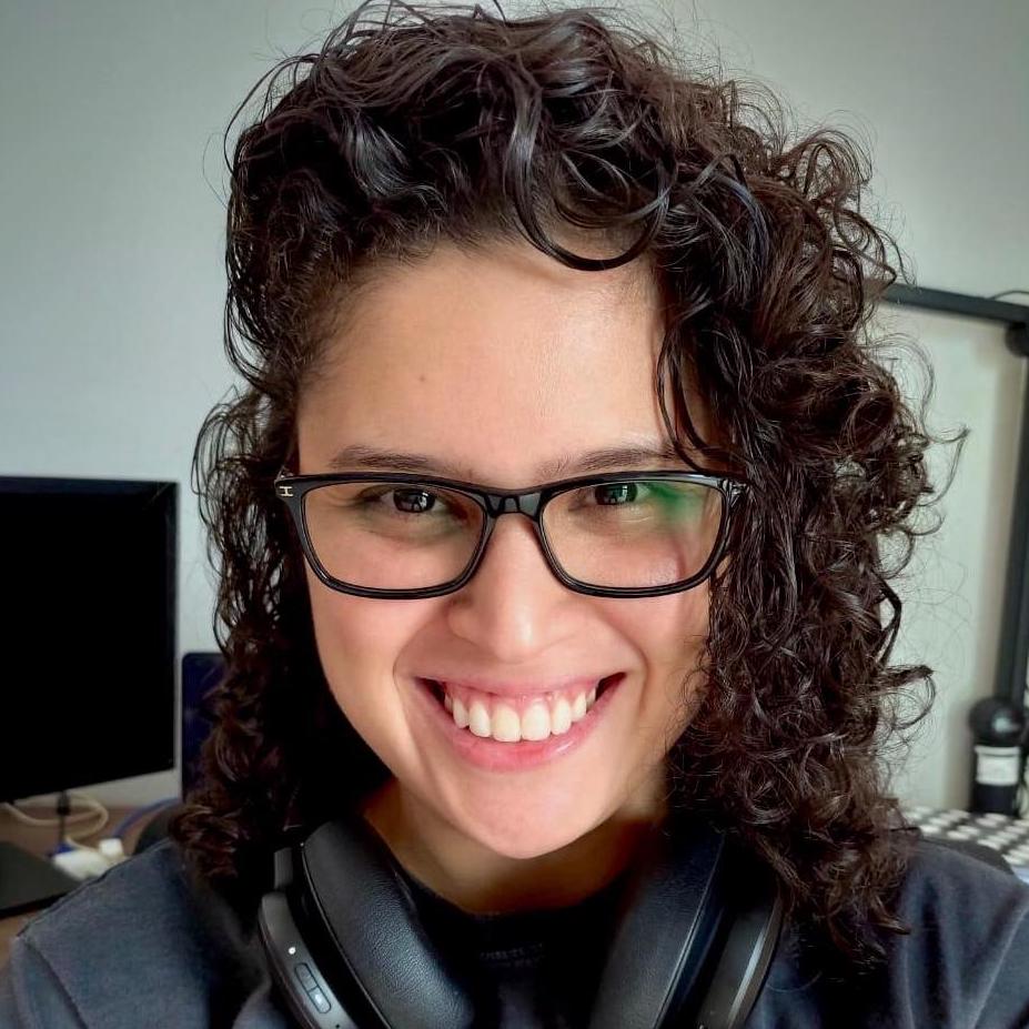 Mulher branca sorridente utilizando óculos preto e caso preto, no fundo tem um monitor de computador.