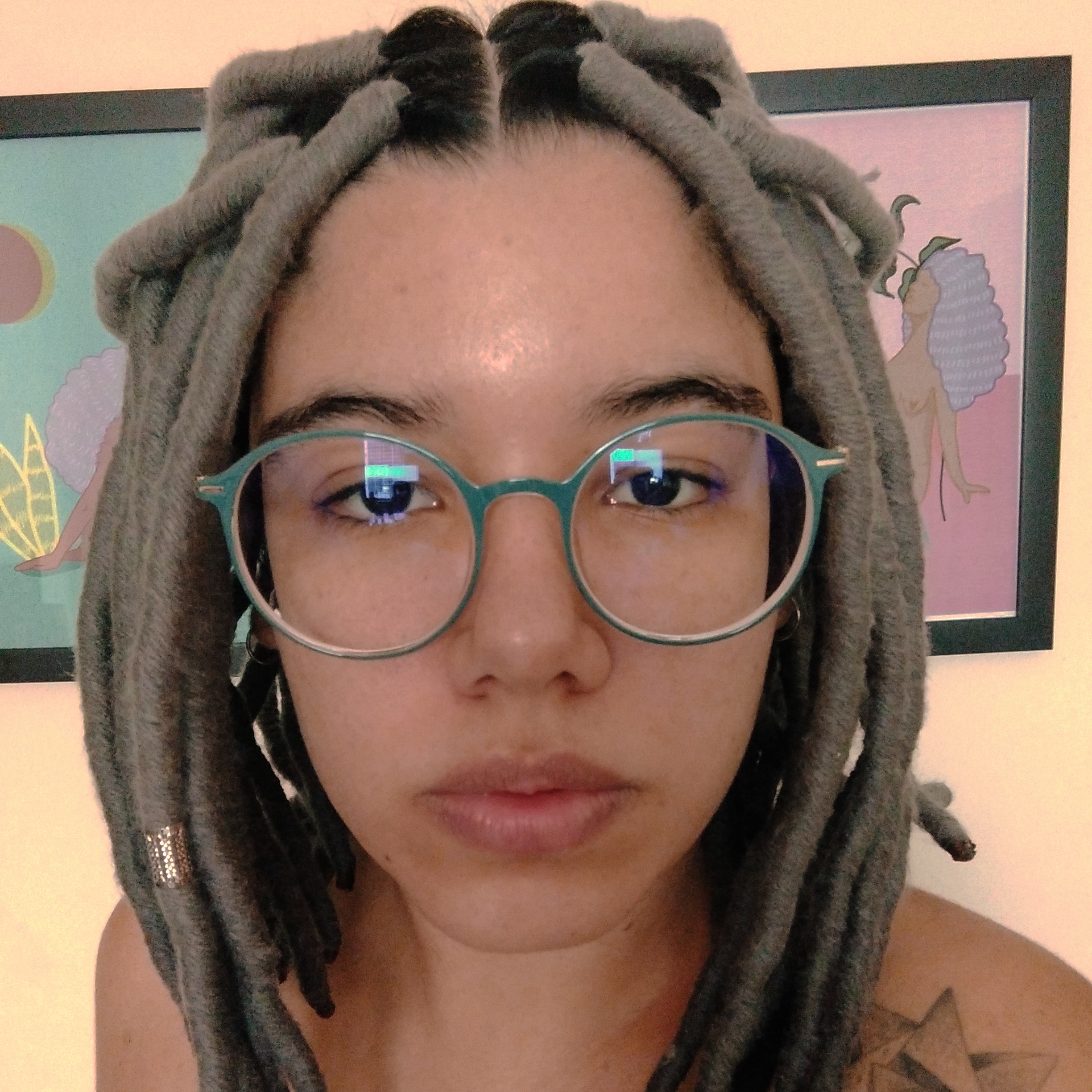 Mulher parda, de dreadlocks cinzas, utilizando óculos preto, no fundo tem dois quadros.